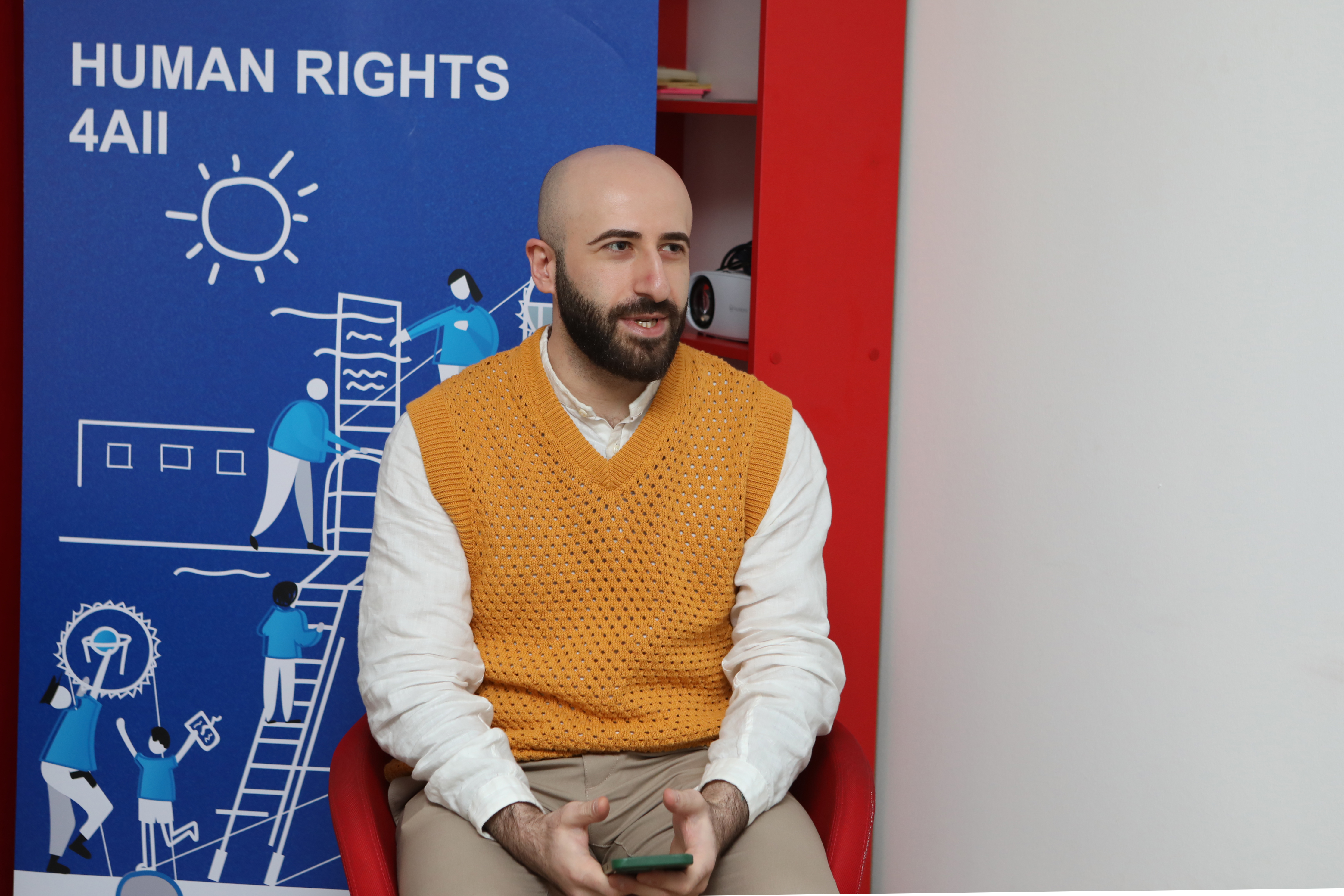 Beka Gabadadze in an orange sweater and white shirt speaking with a blue UNDP banner behind him that reads "Human Rights 4 All"