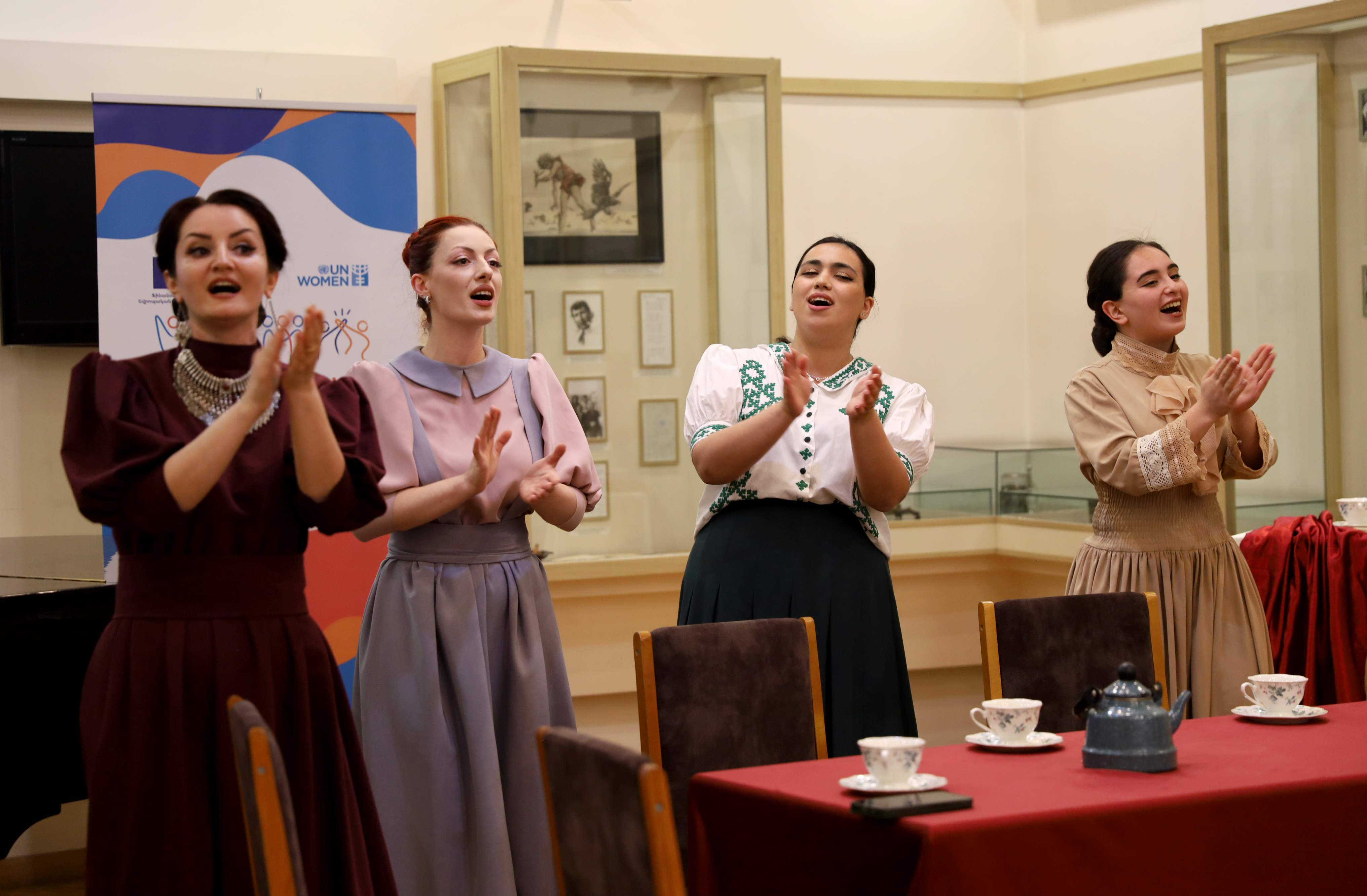 Photo shows four women in traditional clothing singing and clapping their hands