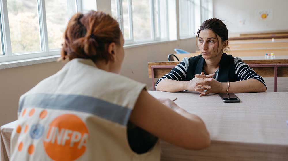 A woman with dark hair sitting at a table and looking into the distance. Another woman wearing a UNFPA vest is facing her.