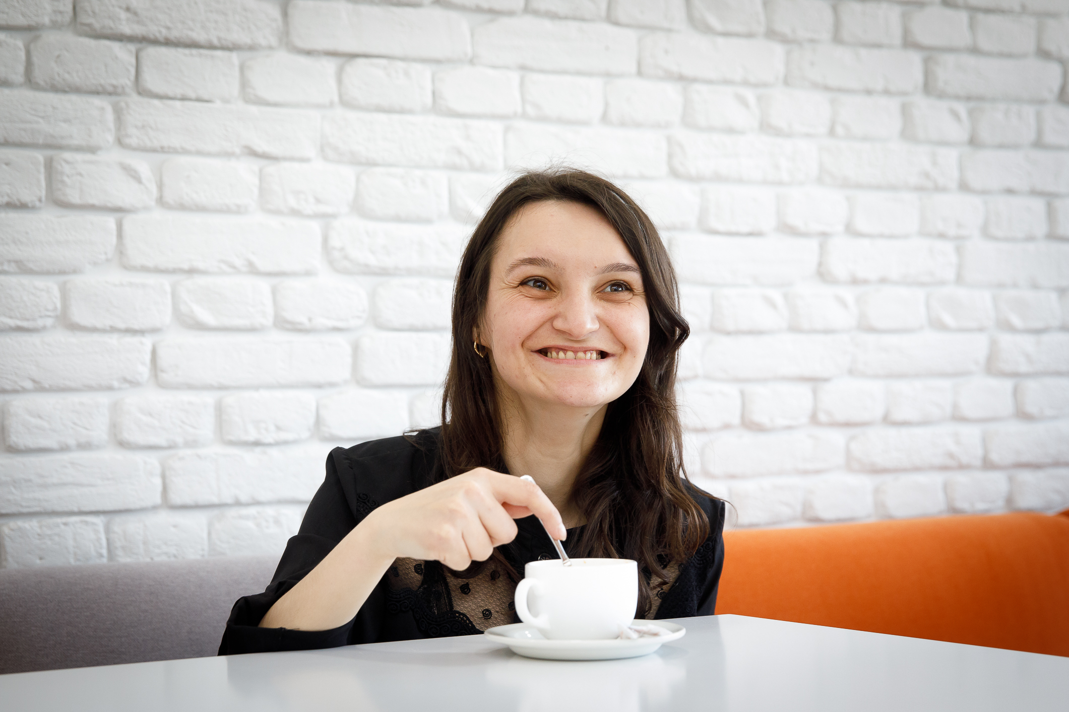A woman in a black shirt sits at a table with a cup of tea, smiling.