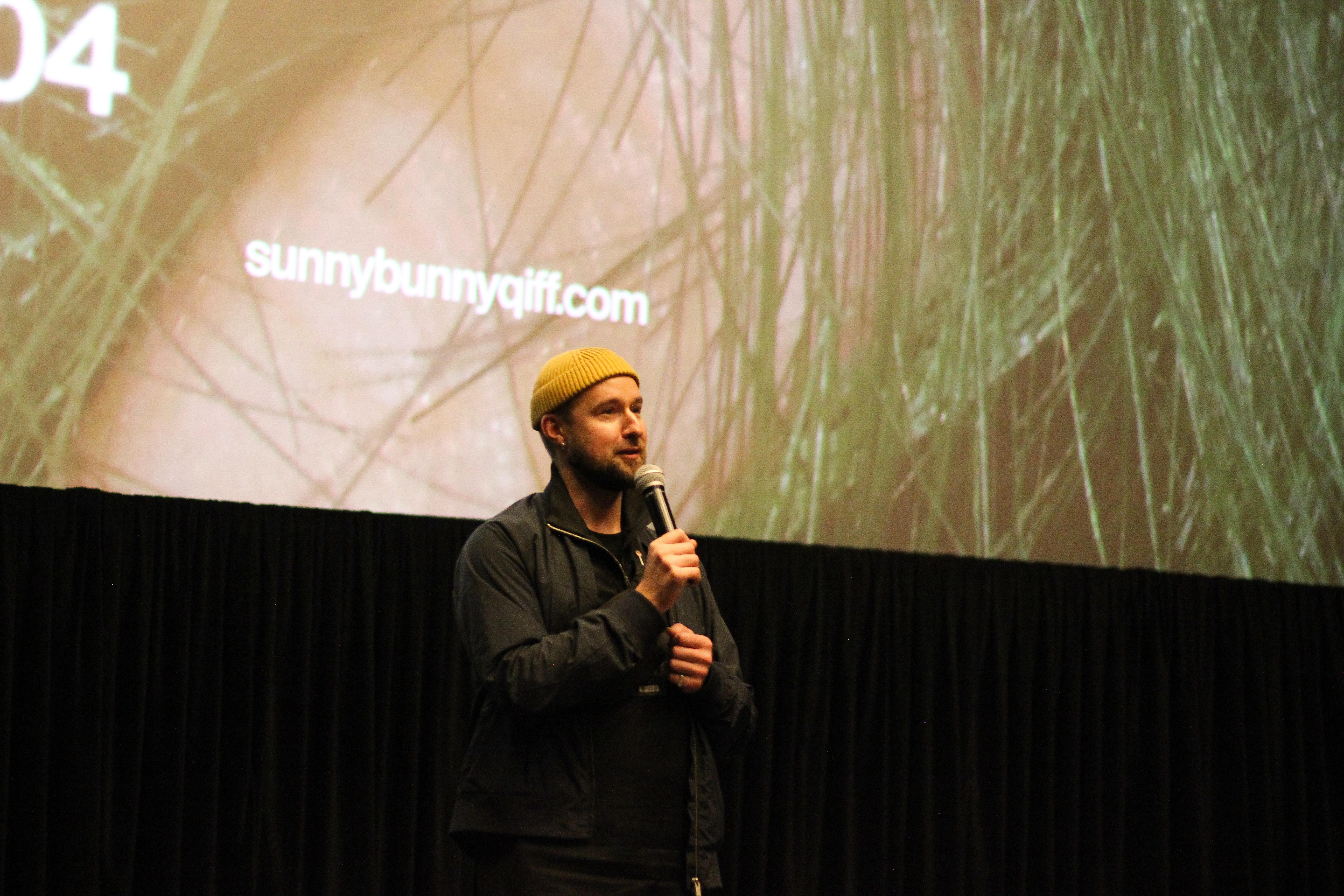 Bohdan Zhuk in yellow beanie stands in front of large movie screen, holding microphone facing unseen audience