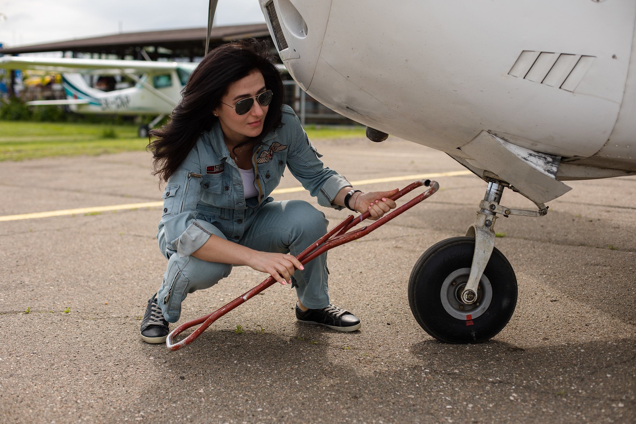 Woman with dark hair and sunglasses kneels down holding a red metal tool beside a light aircraft