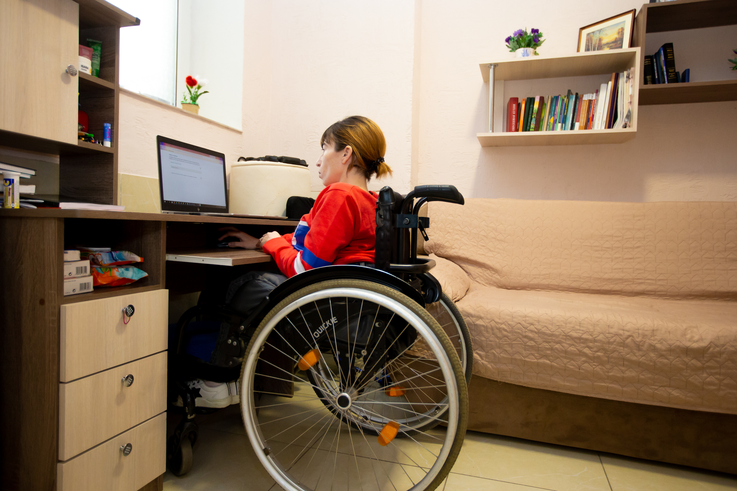 A woman wearing a red shirt and using a wheelchair sits at a desk in her home.