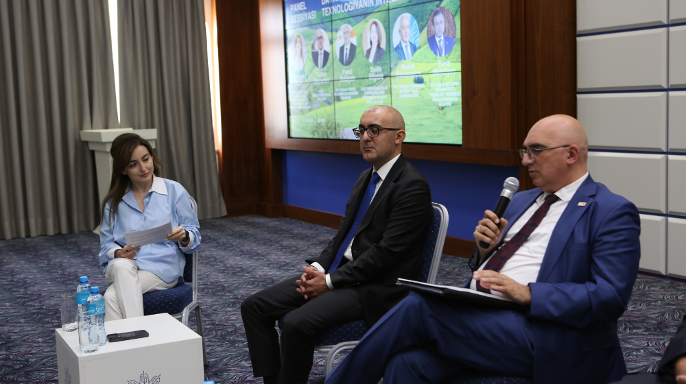 A man in glasses and a blue suit sits in a chair in a conference room speaking into a microphone. A man and a woman also in business dress sit to his right listening. A projector screen behind them shows a list of speakers at the event.