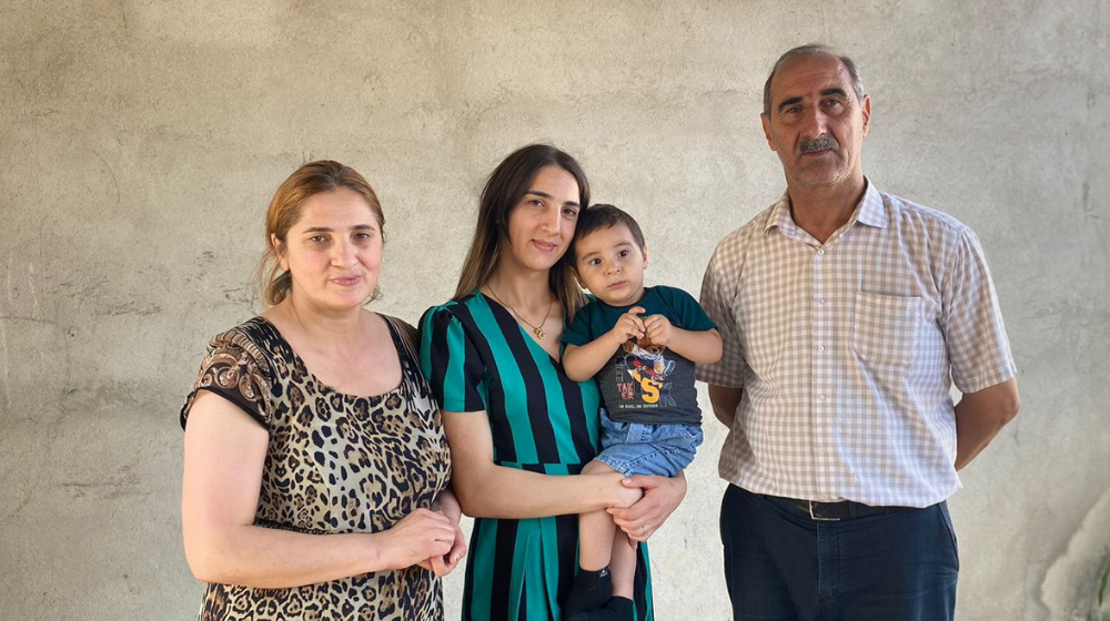 Young woman with long dark brown hair and wearing a green and black striped dress holds toddler boy in her arms. On her right is an older man with a grey moustache wearing a white and beige check-patterned short-sleeve button-down shirt. On her left is a woman with dyed blonde hair wearing a leopard-print dress