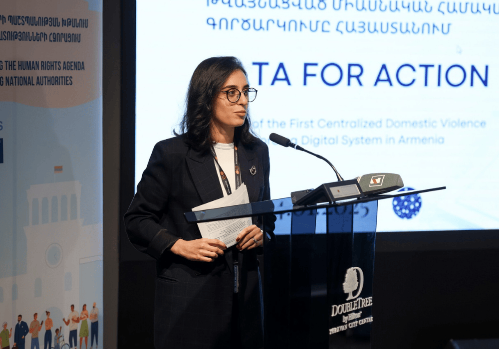 Woman with dark hair and glasses in a dark business suit speaks at a conference room podium holding papers in her hands. A projector screen and banner are behind her