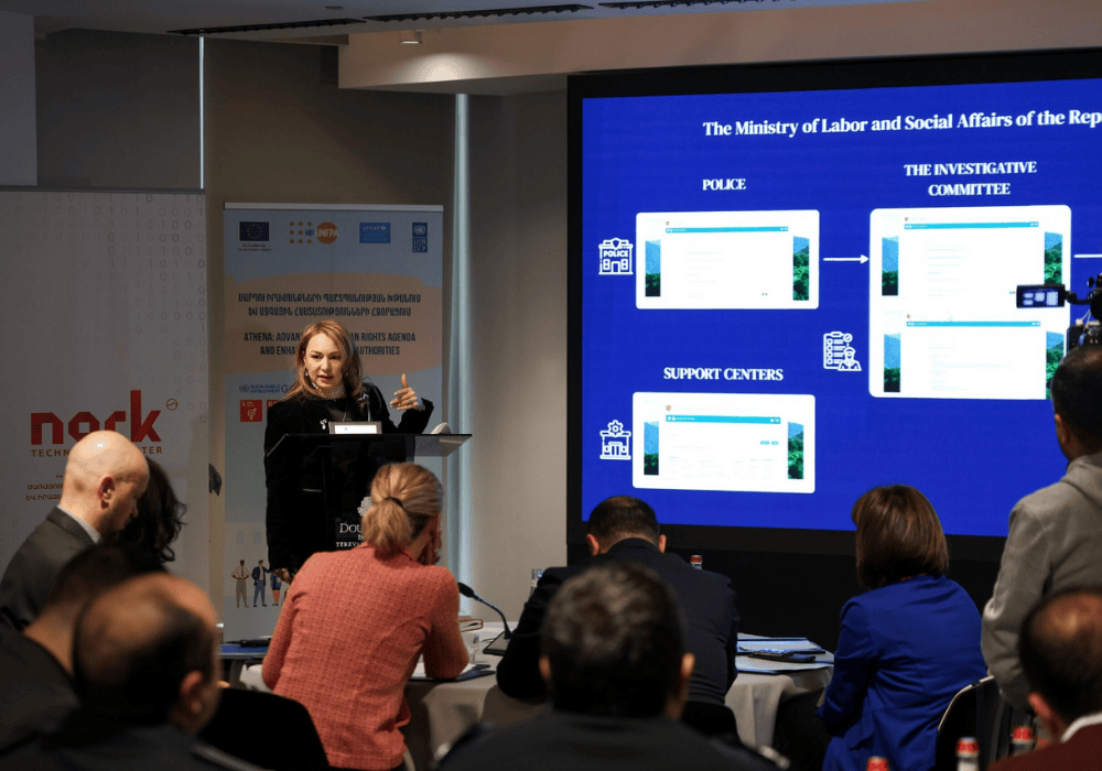 Wide shot of a conference room listening to a presentation by a woman in a black suit and blonde hair. She is standing at a podium speaking next to a projector screen showing a flow chart.