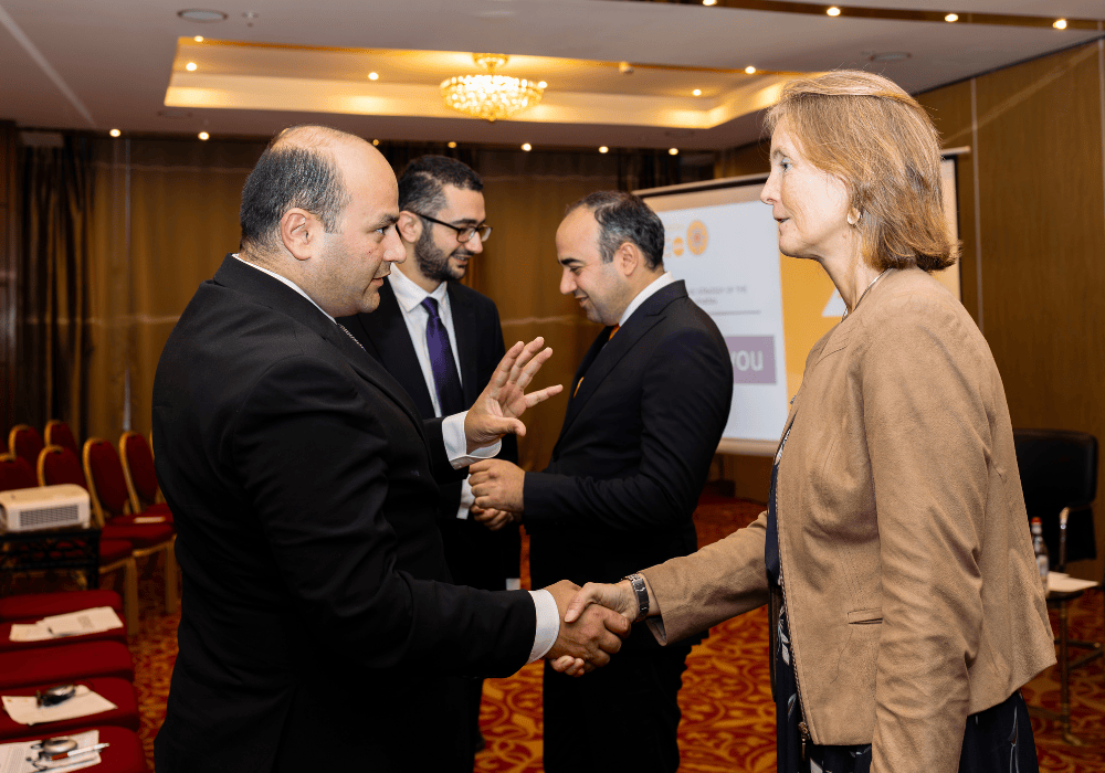 A middle-aged woman with sandy shoulder-length hair and a light brown jacket stands in profile on the right side. She is shaking hands with a bald man in a dark suit who is gesturing with his left hand and appears to be speaking to her. Behind him, two other men in dark suits are speaking to each other smiling, one with a beard and dark rimmed glasses, the other is older and clean-shaven. They are all standing in a hotel conference room with a projector presentation in the background