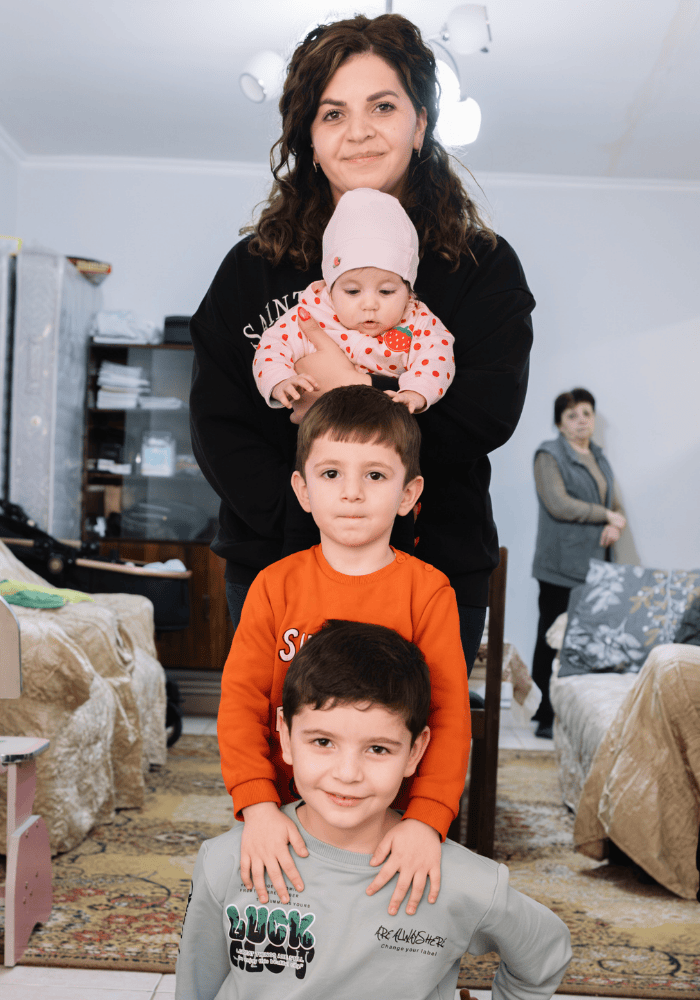 Portrait of a mother and her three young children all in a vertical line. She is standing at the back, has dark brown shoulder-length wavy hair and is wearing all black clothing. She is holding with both arms an infant in a light pink toque and pink shirt with strawberry design. The infant is looking down and reaching toward her brother who is standing below her. He has short brown hair and is wearing an orange long-sleeve shirt. He is staring directly at the camera with a neutral expression. His arms are resting gently on his older brother who is kneeling in front of him. The older boy has darker brown hair and is wearing a light grey tshirt with some stylized text on the chest. He is smiling at the camera. In the background is a family living room with two long sofas along the edges and an older woman standing out of focus in the right corner. 