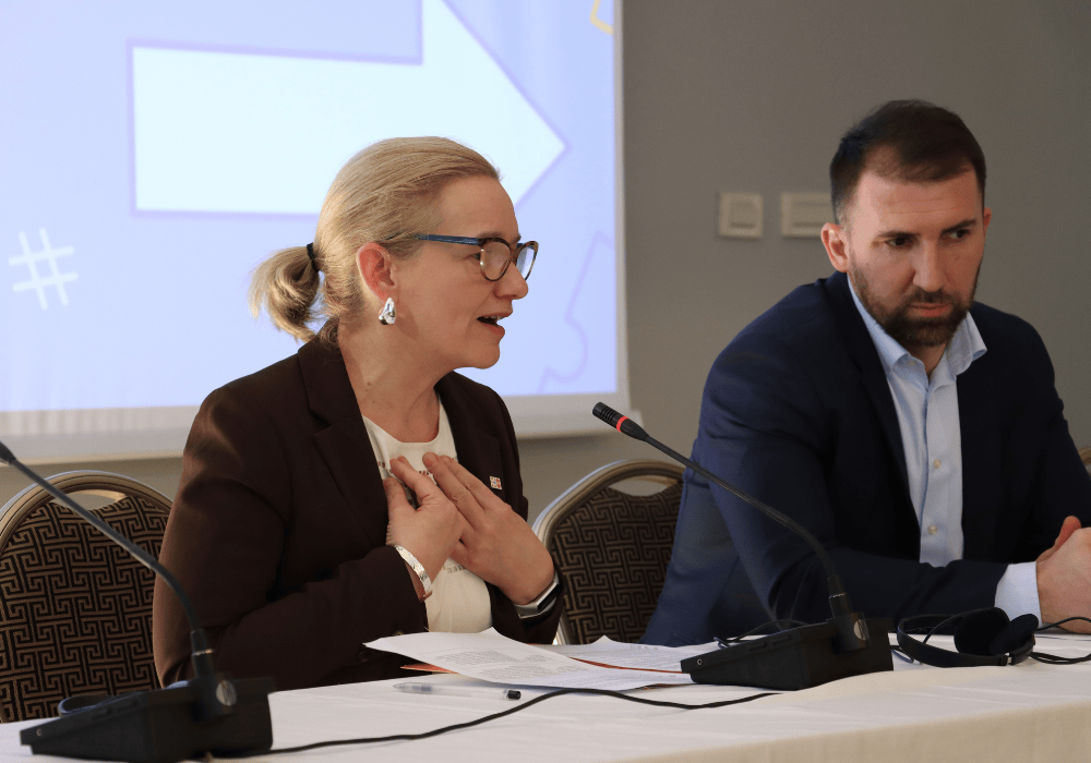 At a press conference in a hotel conference room, a middle-aged woman in a dark business suit with light blonde hair and glasses speaks into a microphone, her arms gesturing towards herself, hands held over her chest. A bearded man sitting to her left, wearing a navy suit, looks downward with a neutral expression