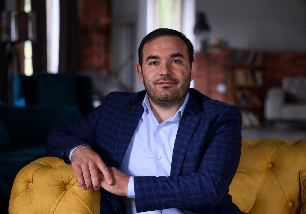 Portrait of a man in a blue checked blazer and light blue collared shirt sitting on a yellow couch with his right elbow resting on the armrest and his hands loosely clasped 