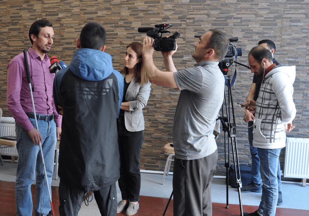 Scene of TV news crews interviewing a visually-impaired man. On the left, a man with brown hair wearing a light purple button-down shirt and jeans holding a blind walking stick. In front of him,  a female journalist with dyed auburn hair in a light grey blazer and a man seen from behiind wearing a long blue-black Adidas track jacket are holding up microphones to the visually impaired man's face. Behind them, three more men are standing to film the interview. One an in a grey polo shirt is holding the camera up in his hands. Another in a white hooded zip sweatshirt is standing behind a larger camera on a tripod with white headphones plugged in, he is staring down at his phone. The last man is standing against the wall, barely visible behind the others. 