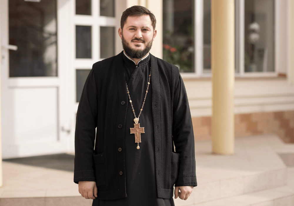 Portrait of Moldovan priest Andrei Apetrei, dressed in a black religious robe, with a black button-up vest and a large gold cross around his neck. 