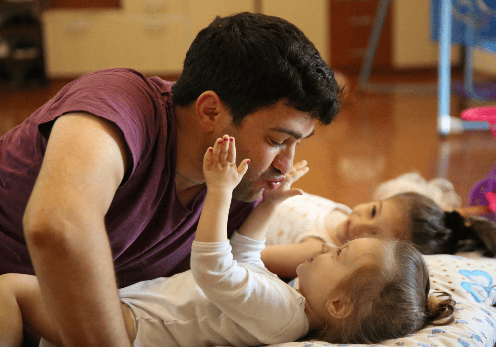 A father plays with his two young daughters on the ground: Man with dark hair wearing a mauve tshirt leans over an infant girl making a face with puckered lips. Two infant girls smile at him, one in the foreground in a white onesie lying on a thin pillow with butterfly designs holds the man's face with two hands, the other girl in the background also on her back smiles at him.