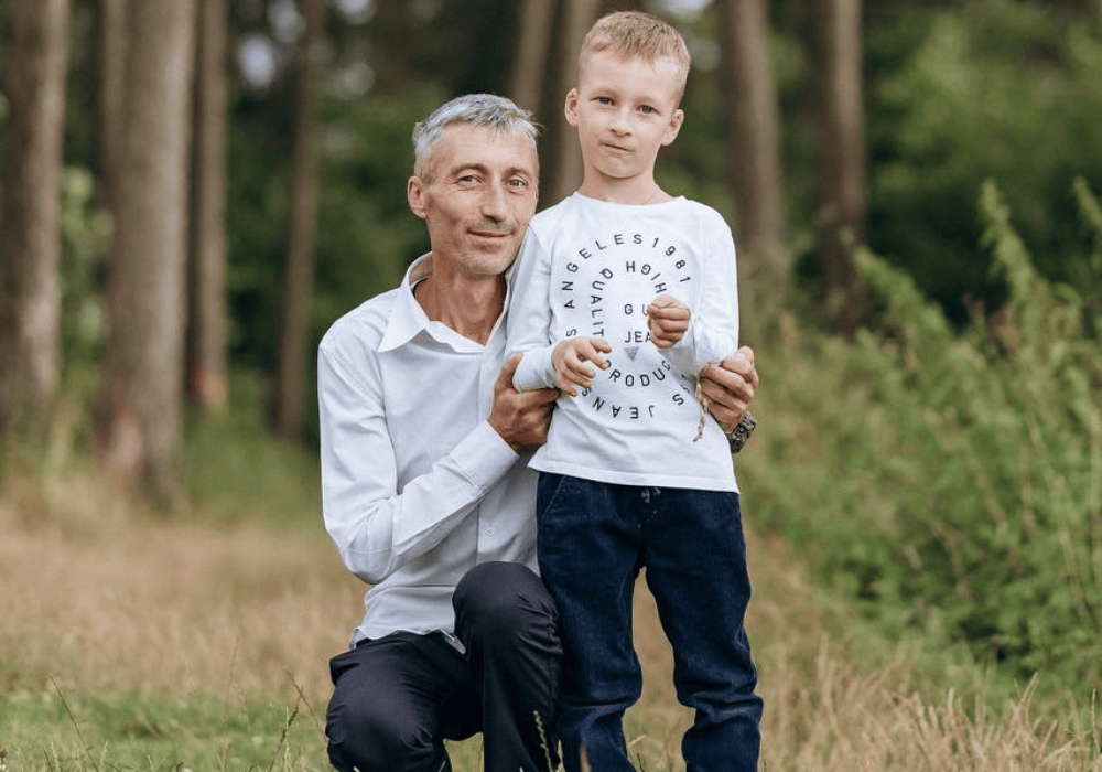 Portrait of a father and son standing in the woods: On the left, an older man with grey hair and a white button down shirt and jeans kneels next to a young boy with short blonde hair who is also wearing jeans and a white shirt with text swirled across the front. The boy's arms are bent loosely in front of him, the older man's arms are wrapped gently around the boy. They are both looking directly at the camera with soft closed-mouth smiles