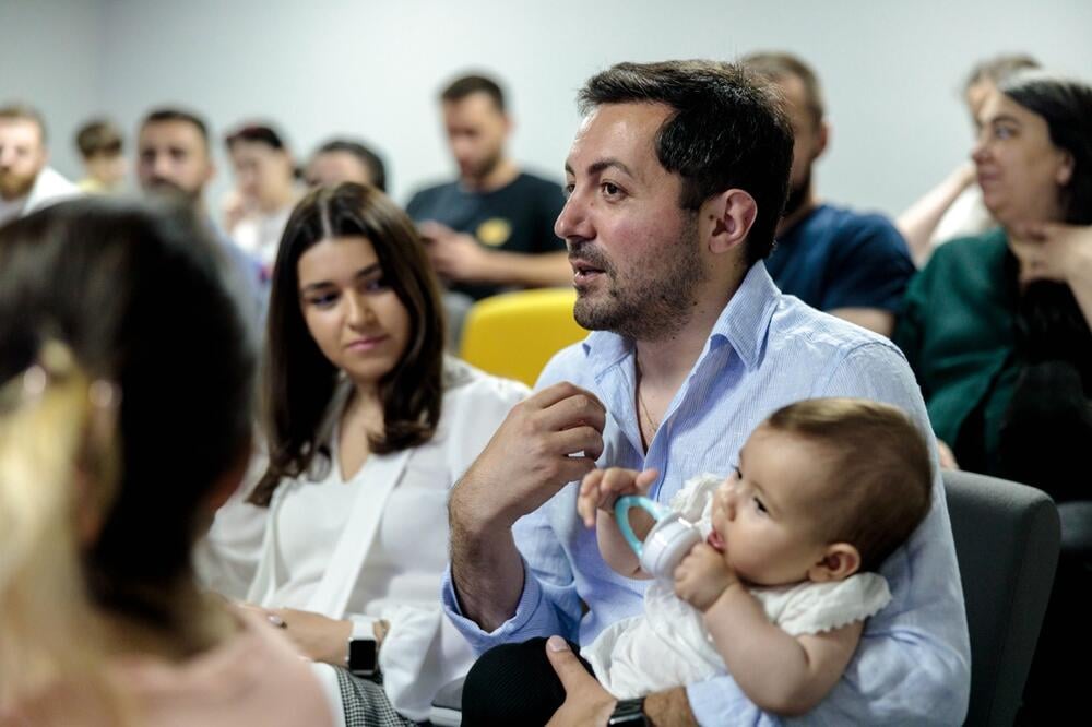 A man with dark hair is holding a baby and speaking with animation in a crowd.