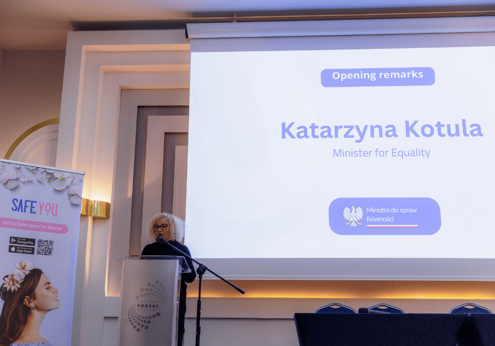 Woman with curly blonde hair and large black-framed glasses wearing a black turtleneck stands at podium in conference room. Projector screen to her right reads "Opening remarks / Katarzyna Kotula / Minister for Equality". On her left is a banner with the SafeYou logo and the image of a woman tilting her head up and smiling with eyes closed. 