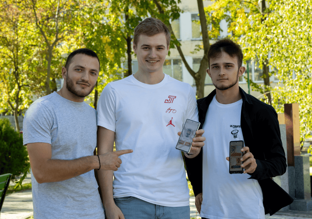 Three young men stand in a line and smile at the camera. The two on the right -- one with dark hair and beard and black jacket over a white shirt on the right; a taller blonde man in a white tshirt in the centre -- hold up their phones with the AIDor app on the screen. On their left, a man with short dark hair and beard with a light-grey tshirt points to their screens and gives a closed-mouth smile. 