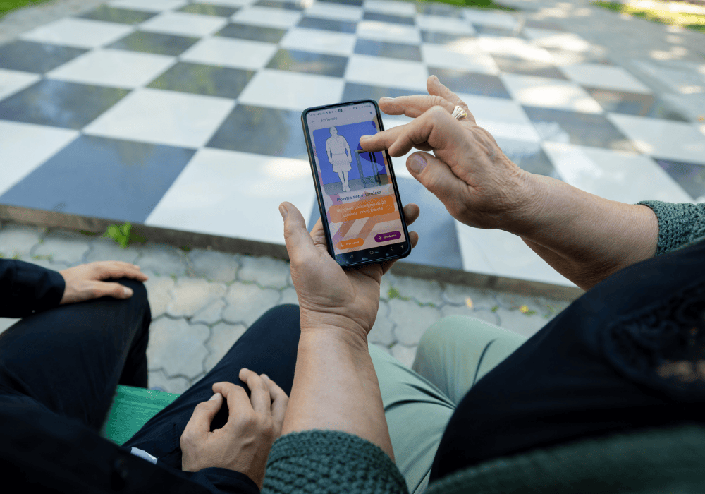 Camera angle looking over the shoulder of two people sitting next to each other on a park bench, only their legs and arms are visible, not their faces. Woman on the right is holding a smartphone device and scrolling on an image from the AIDor app of an illustrated human body. 