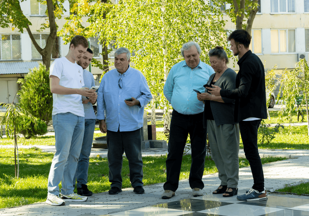 Full-body photo of six people standing together in an outdoor park. Two older men in light blue button-down shirts and one older woman in a light green cardigan stand in the middle. On the left, two young men demonstrate something on a white smartphone while one of the older men looks over their shoulder. On the right, a third young man in a black jacket holds out his smartphone next to the woman, who is also holding her own phone and looking at the screen. Behind them are some leafy trees and an apartment block.