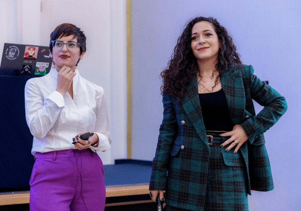 Woman with dark curly hair in a green checkered suit stands looking to the distance with a hopeful look. To her left is a woman with short dark hair and glasses in a white collared shirt and bright purple trousers, holding her chin and smiling thoughtfully in the same direction