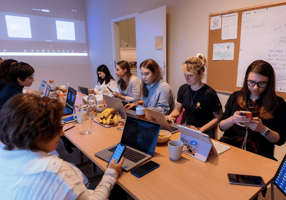 Group of women gather around a meeting room table, each one holding their own phone or laptop. On a projector screen at the front is SafeYOU's training presentation