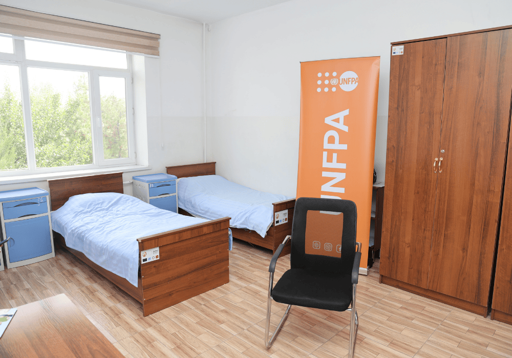 Room for GBV survivors: On the back wall, large set of windows looking on to trees. Under the window sill are two single beds with dark wooden bed frames, each one with a light blue-and-white bedside table to the left. On the right side wall, a large dark wooden dresser. A black chair sits empty in the middle of the room. An orange UNFPA banner behind it. 