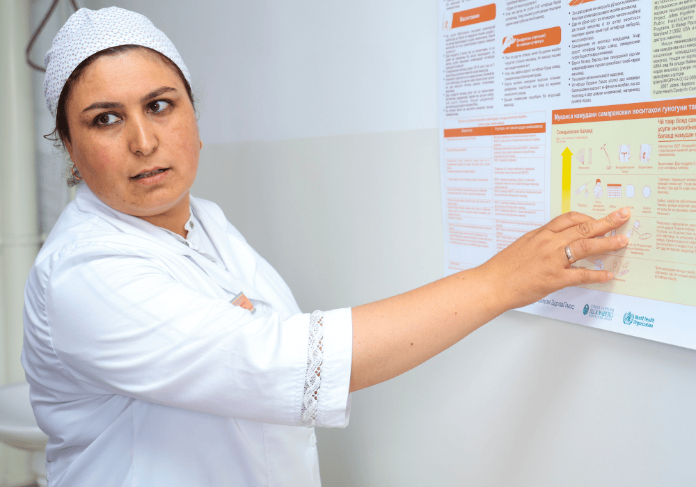 Close up image of woman in doctor's short-sleeved white coat and cap pointing to an information poster on the wall. She has dark brown eyes and eyebrows with a neutral expression as she looks to the left off camera. 