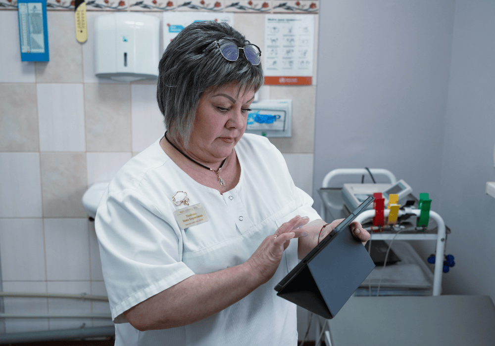 Nurse Inna in the clinic room looking at an electronic tablet. She is wearing a white hospital uniform, her glasses are on her head.