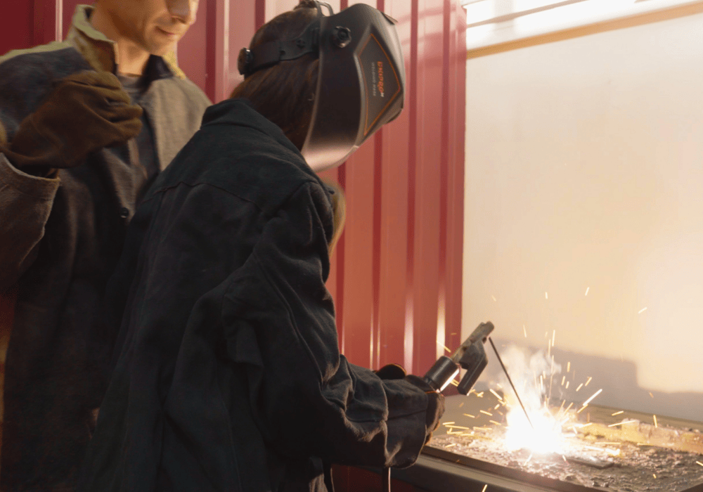 Woman with brown hair in a welder's helmet uses a welding torch at an industrial looking workbench with sparks flying. Behind her, slightly out of focus and only visible up to the nose, a man stands observing in a thick grey coat and brown gloves