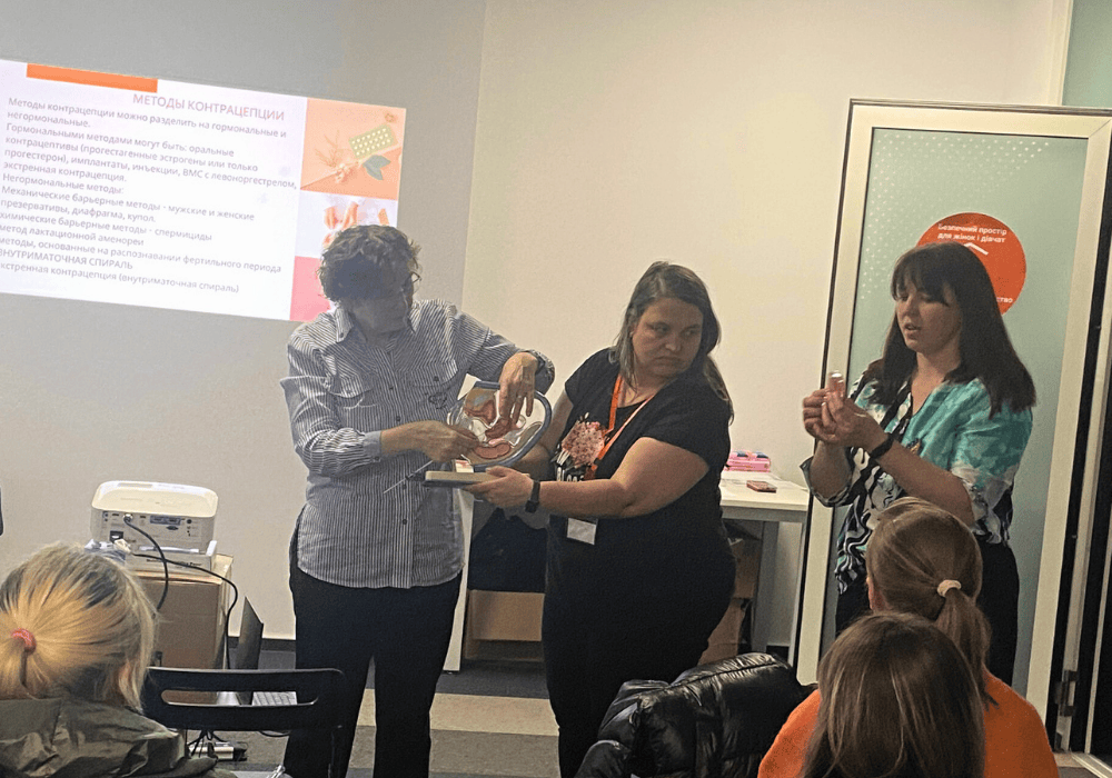 Three women stand in front of a room full of women giving presentation on sexual and reproductive health. The two women on the left hold up a model of the female reproductive organs, while the woman on the right gives an explanation holding a smaller piece. Behind them, a Power Point presentation is projected on to the wall