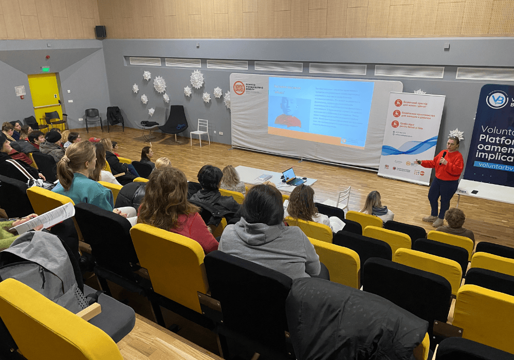 An auditorium full of women listen as a woman in a red sweater and black jeans stands at the front holding a microphone giving a presentation. Projector screen beside her shows information about psychosocial support in Ukrainian. Banner with UNFPA Safe Spaces logo and information is in the background.