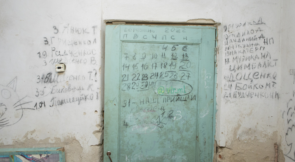 Close-up shot of words handwritten in black on a white wall with a calendar marking the days spend in the basement sketched on a wooden mint-coloured door