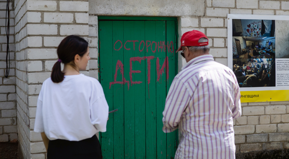 Older man wearing a red cap and a young woman with dark hair in a ponytail stand in front of green wooden door with red graffiti showing entrance to school basement