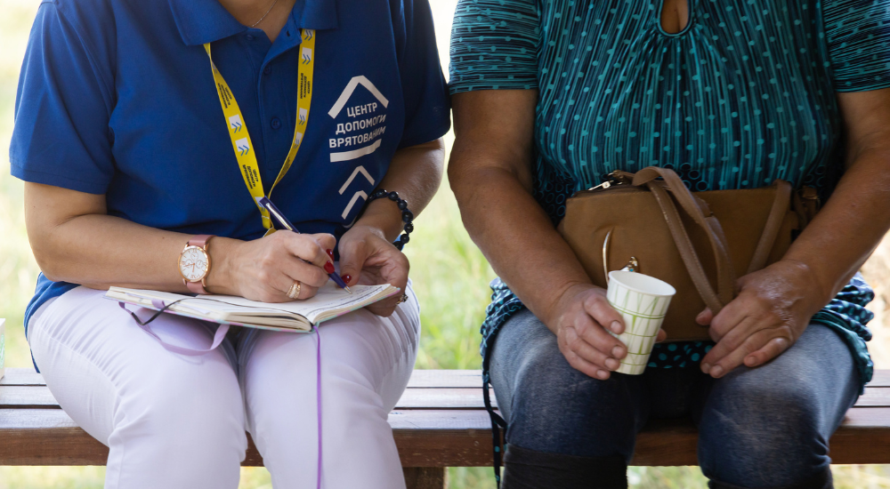 Close-up shot of two women sitting next to each other, faces not visible, one in a blue UNFPA shirt and yellow lanyard taking notes, while the other holds a paper cup with her brown purse in her lap 