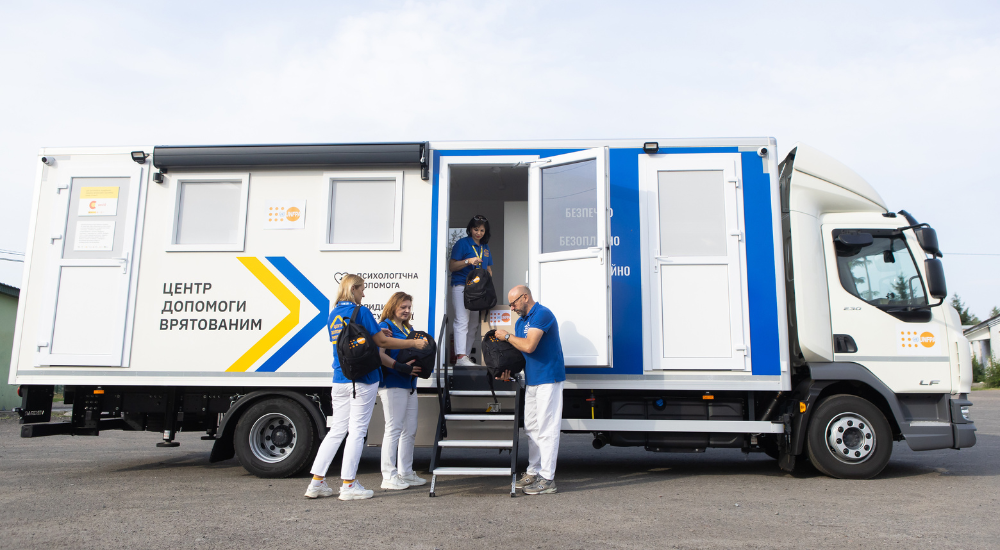 Four UNFPA volunteers in blue shirts prepare black backpacks outside their white mobile support centre truck