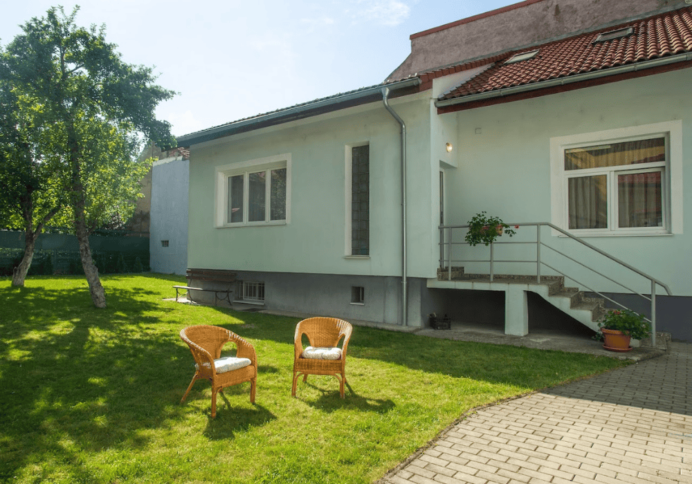 Shows exterior of Agapedia Foundation office. A one-story mint-coloured building with a set of stairs leading to the main entrance. There is a large grass yard with two empty chairs and trees in the background