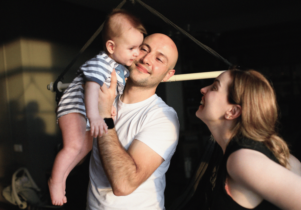 Portrait of a young family. Father is standing in the centre facing the camera, wearing a white tshirt, and holding a baby up in the air with both hands, smiling lovingly at the child. The baby is in a white and blue striped romper and is smiling. On their right, the mother looks happily up to the child, smiling widely. She has light brown hair pulled into a loose ponytail and is wearing a black sleeveless shirt. 