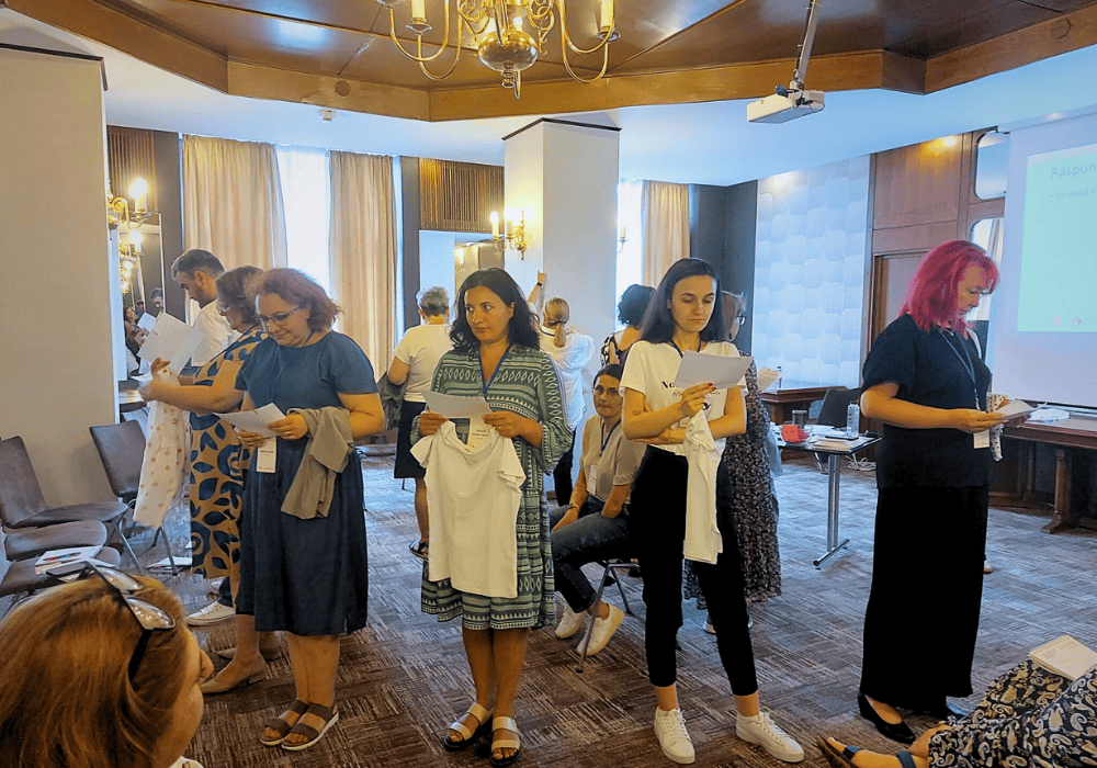 A group of women participate in a training exercise where they stand in a circle with their backs facing the woman sitting in the centre. The standing women are holding papers and shirts. The activity is being held in a hotel conference room.