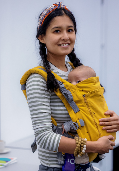 Portrait of a young woman with dark hair in pigtails and a red patterned hair band. On her front, she is cradling a yellow baby carrier with her infant peeking out from the top. 