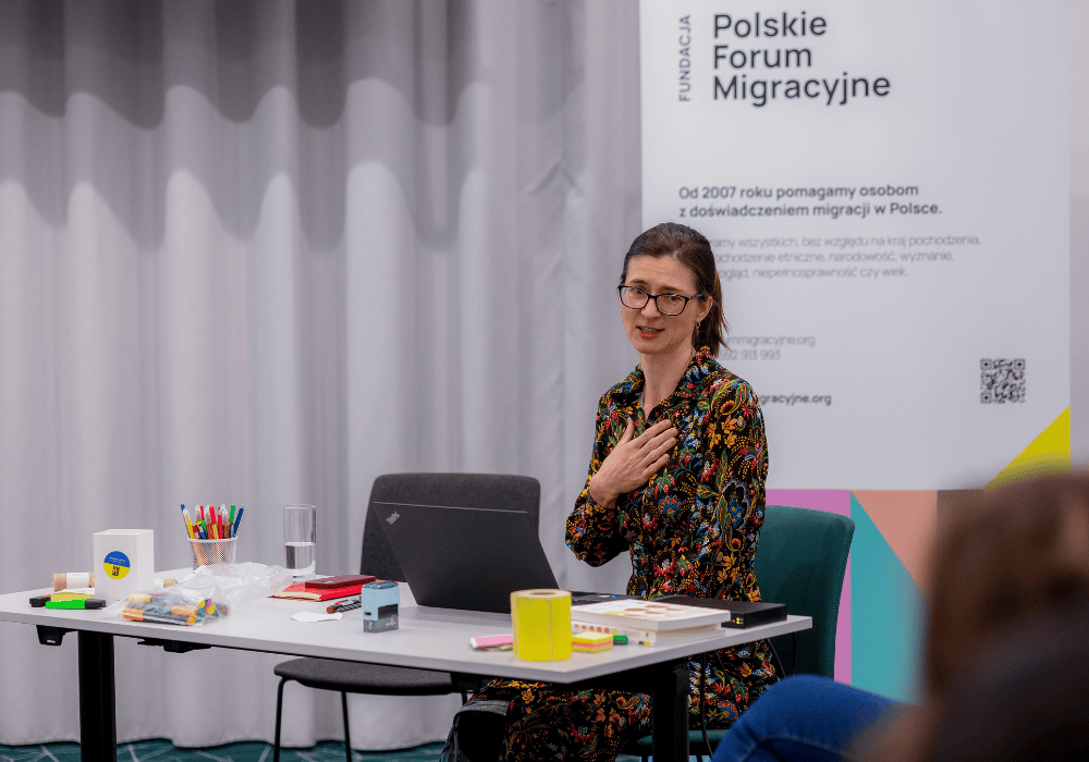 Woman with dark brown hair, wearing black-framed glasses and a floral-print dress, sits at a table while speaking to an audience. She holds her right hand over her heart. There is a variety of office stationery on the desk in front of her. Behind her on the right is a Polish Migration Forum banner. 