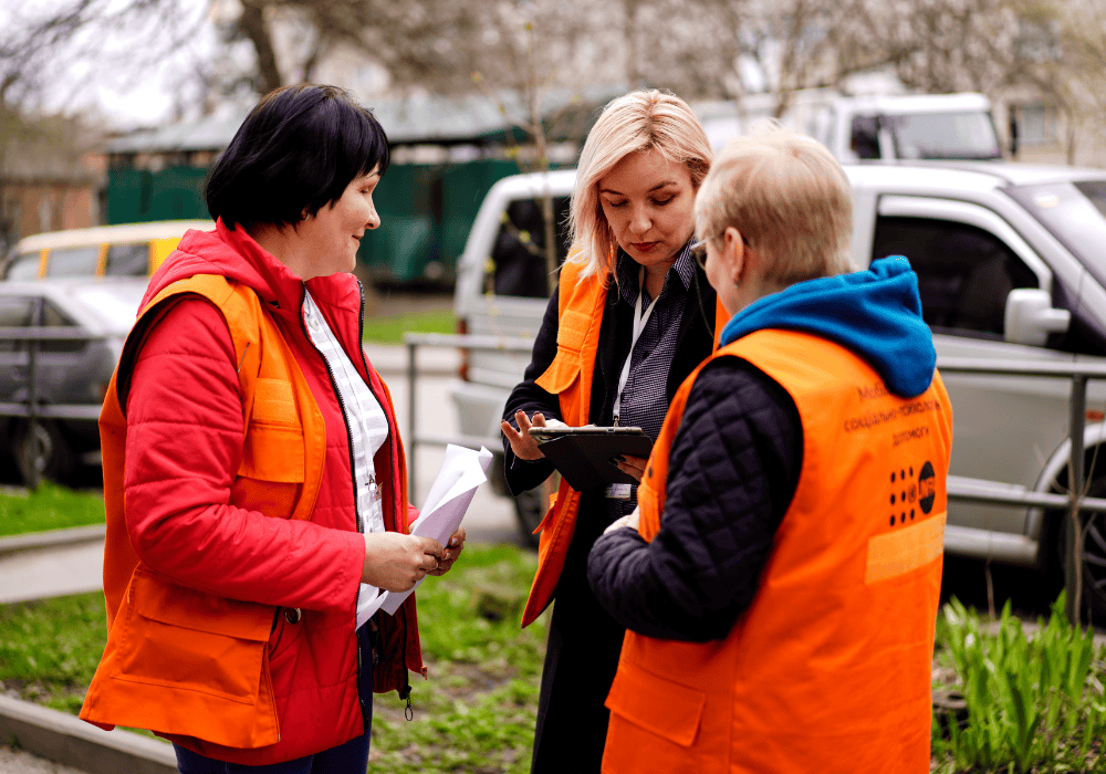 Three women in neon orange UNFPA mobile support team vests stand in a circle together outside. In the middle is a woman with blonde hair looking down at a clipboard. 