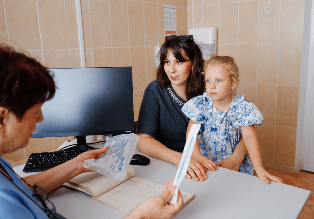 Inside a clinic room, a woman with dark brown hair and dark tshirt sits at a desk speaking to a healthcare worker while holding her young daughter in her lap. The daughter has blonde hair and is wearing a blue and white dress. The healthcare worker sitting across from desk is wearing blue scrubs and is holding a medical implement in her hands, explaining to the other woman. There is a computer the desk. 