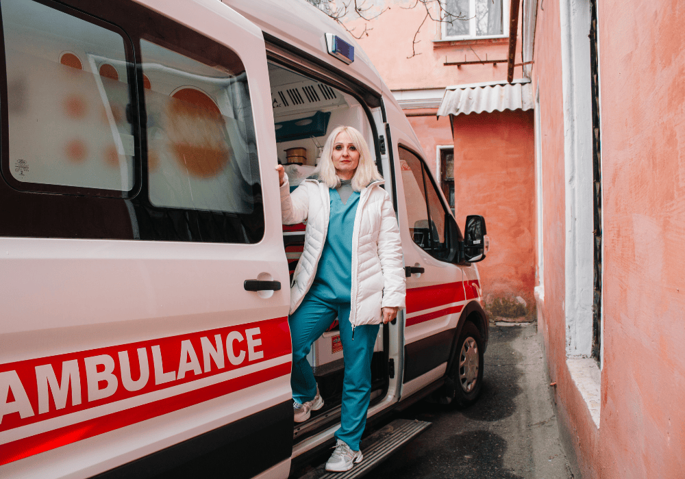 Ambulance with UNFPA and AECID logos on its side is parked outside a salmon-coloured building. A woman in standing beside the ambulance looking directly at the camera with a neutral expression.  She has shoulder-length white hair, and is wearing medical scrubs and a white puffy winter coat, her hands tucked in the coat pockets