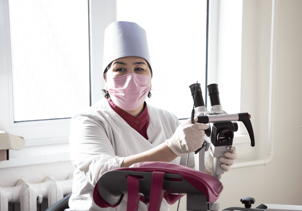 Female health worker sits in a clinic exam room. She is wearing a white lab coat and toque with a pink face mask and latex gloves. In front of her is a microscope for conducting cervical screenings. The gynaecological exam table is out of frame but one of the stirrups is in the bottom right corner, below the woman's arm