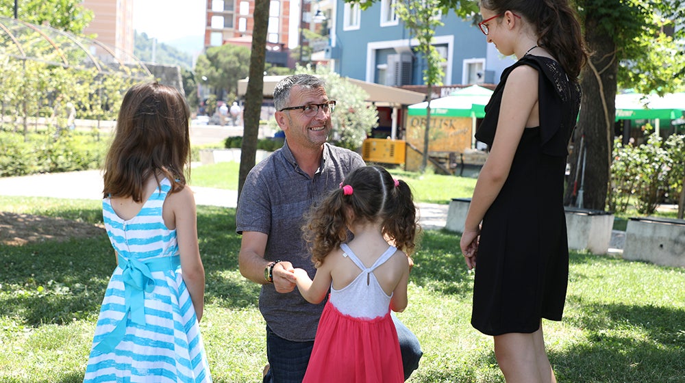 A father kneels in the grass while playing with his three daughters