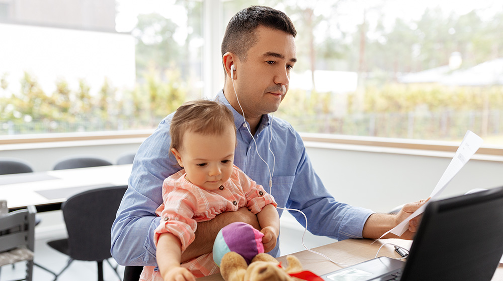 A man in a blue shirt sits at a desk. He is holding a baby girl and looking at a stack of papers in his hand.
