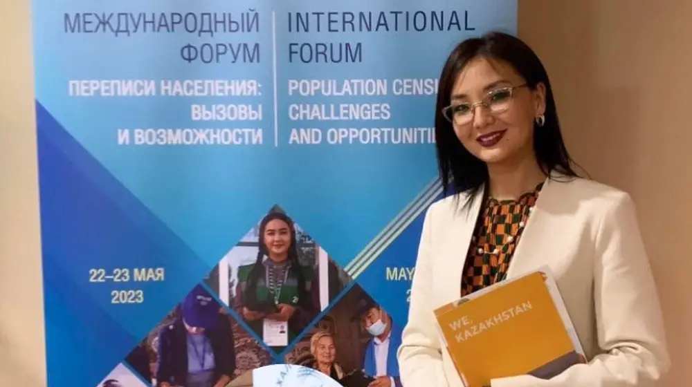 A woman is standing in front of a banner. She is wearing glasses and smiling and she is holding a folder.