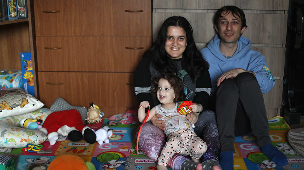 A woman, man, and girl child sit on the floor of a house. All three are smiling and are surrounded by toys and household items.
