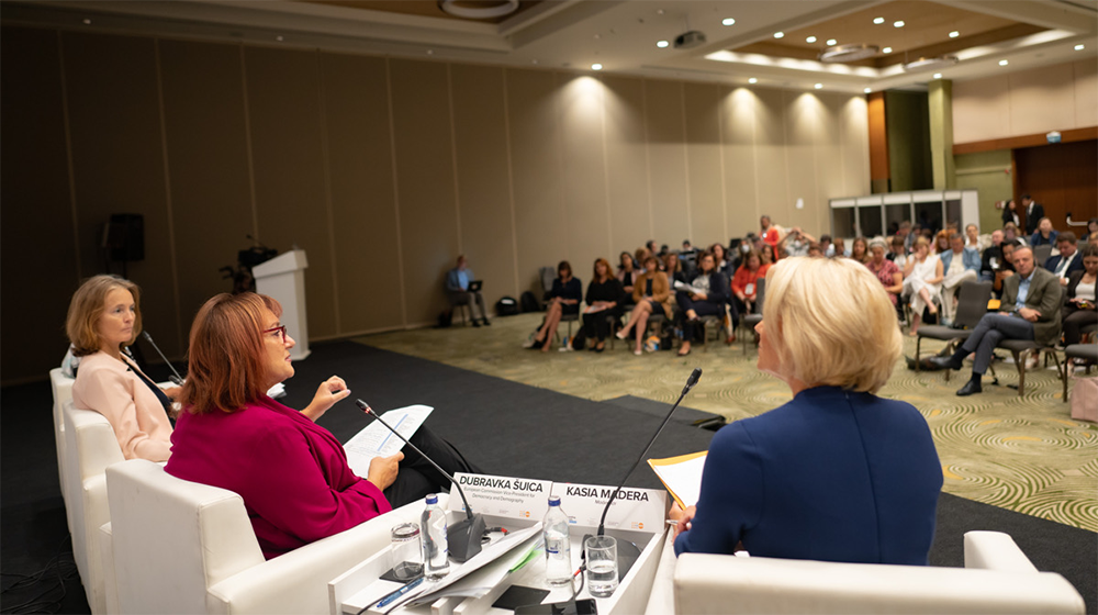 Three women sit in white chairs with their backs to the camera, having a conversation. Behind them, a crowd of people watches.